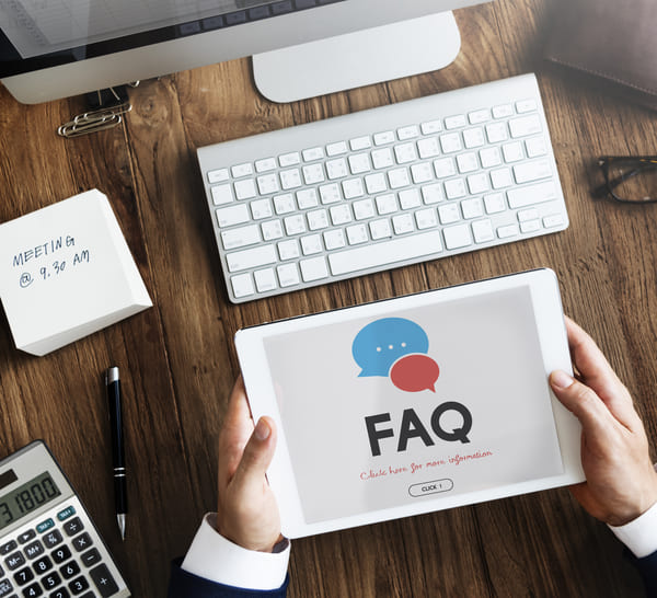 Une assistante de direction indépendante tient une tablette affichant une page FAQ (Foire Aux Questions), illustrée par des bulles de dialogue. La scène se déroule sur un bureau organisé avec clavier, calculatrice, stylo, et bloc-notes indiquant un rendez-vous. Cette image évoque les services d’assistanat, le soutien administratif clé et l’organisation professionnelle à distance.