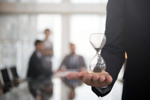 Un homme en costume tenant un sablier dans sa main, illustrant la notion de temps précieux en entreprise. En arrière-plan, des collaborateurs flous discutent en réunion. Cette image évoque l’importance de déléguer les tâches administratives pour gagner du temps et se concentrer sur l’essentiel, un service offert par une assistante de direction indépendante.